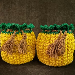 May include: Two crocheted pineapple-shaped pouches with green tops and brown tassels. The pouches are yellow and have a textured surface.