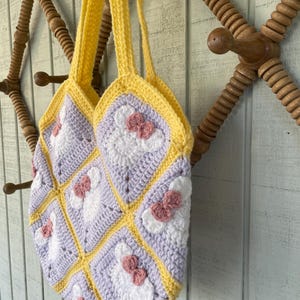 May include: A crocheted tote bag with a yellow and purple patchwork design. Each square features a white crocheted flower with a pink bow.