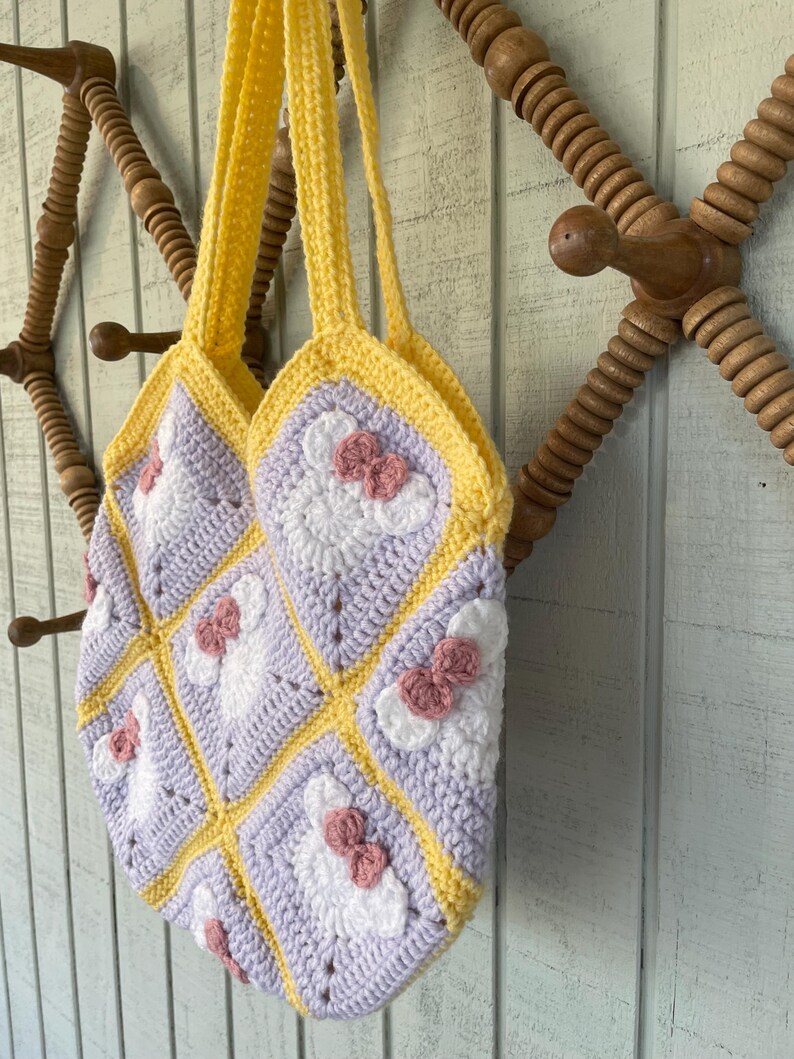 May include: A crocheted tote bag with a yellow and purple patchwork design. Each square features a white crocheted flower with a pink bow.