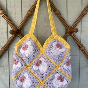 May include: A crocheted tote bag with a yellow and purple geometric pattern. The bag has a white and pink flower design with a bow in the center of each flower.