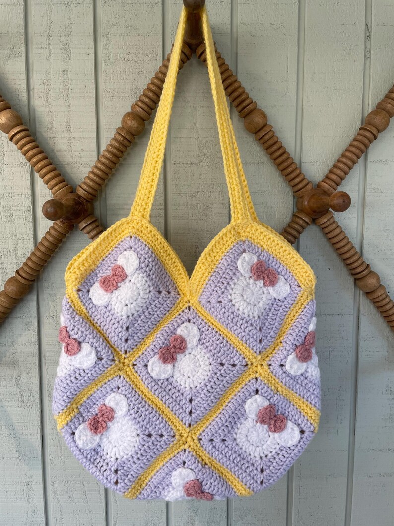 May include: A crocheted tote bag with a yellow and purple geometric pattern. The bag has a white and pink flower design with a bow in the center of each flower.