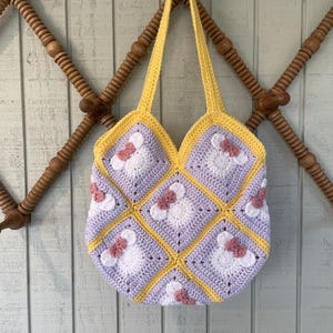 May include: A crocheted tote bag with a patchwork design featuring squares of lavender, white, and yellow yarn. Each square has a pink flower with a white center. The bag has yellow straps.