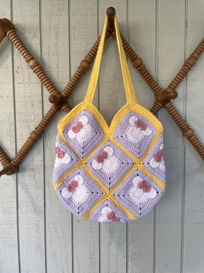 May include: A crocheted tote bag with a patchwork design featuring squares of lavender, white, and yellow yarn. Each square has a pink flower with a white center. The bag has yellow straps.
