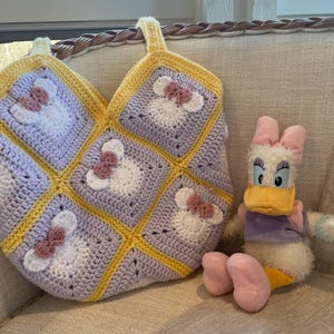 May include: A crocheted tote bag with a yellow and purple color scheme featuring a repeated pattern of a cartoon mouse with a pink bow. The bag is sitting on a light beige couch next to a plush duck toy.