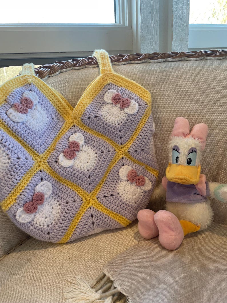 May include: A crocheted tote bag with a yellow and purple color scheme featuring a repeated pattern of a cartoon mouse with a pink bow. The bag is sitting on a light beige couch next to a plush duck toy.