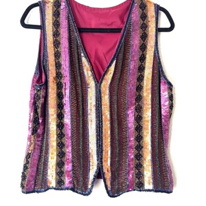 May include: A sleeveless vest with vertical stripes in shades of pink, orange, and brown, embellished with sequins and beads. The vest has a V-neck and is hanging on a black hanger against a white background.
