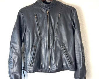 May include: Black leather jacket with a stand-up collar and multiple zippered pockets. The jacket has a front zipper closure and zippers on the sleeves. The back of the jacket has a lace-up detail. The jacket is hanging on a wooden hanger.