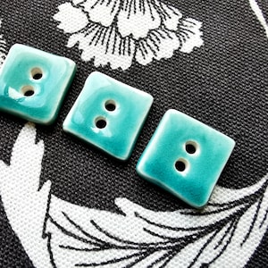 May include: Four square ceramic buttons in a teal blue color with two holes each. The buttons are arranged in a row on a black fabric with a white floral pattern.