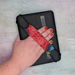 May include: A black Kindle e-reader with a red and patterned hand strap. The strap has a floral design and gradient colors of blue, purple, and yellow. The Kindle is held in a hand.