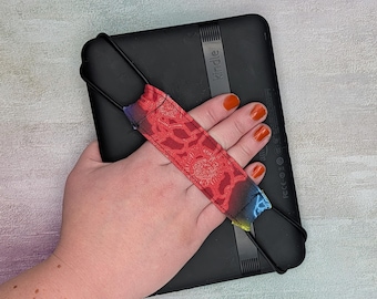 May include: A black Kindle e-reader with a red and patterned hand strap. The strap has a floral design and gradient colors of blue, purple, and yellow. The Kindle is held in a hand.