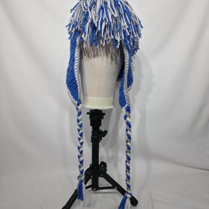May include: A handmade blue, white, and gray crochet hat with long braided ear flaps. The hat features a shaggy top with blue, white, and gray yarn. The ear flaps are braided with blue, white, and gray yarn and end in tassels.