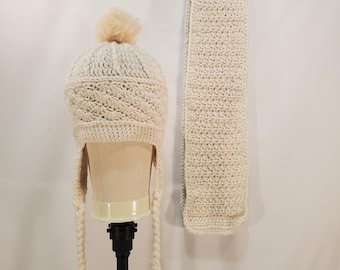 May include: A cream-colored, crocheted hat with ear flaps and a matching scarf. The hat features a textured pattern and a fluffy pom-pom on top. The scarf is long and rectangular, hanging on a black hanger.