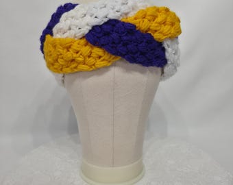 May include: A hand-crocheted headband in a braided design. The headband features a color block pattern with sections of white, purple, and yellow yarn. The headband is displayed on a mannequin head against a white background.