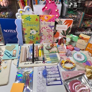 May include: A collection of stationery, beauty products, and accessories. Includes notebooks, pens, a tote bag with the text "Just One More Chapter", face masks, and a variety of other items. The color palette is bright and cheerful.