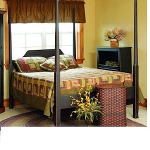 May include: A dark wood four-poster bed with a patterned quilt in a bedroom setting. The quilt features a patchwork design with green, red, and brown squares. A woven basket and a small cabinet are also visible.