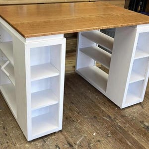 May include: White wooden craft table with a natural wood top and open shelving on both sides. The table has a white painted finish and is on wheels.