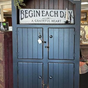 May include: A blue painted wooden cabinet with two doors and a top shelf. The cabinet has a sign on the top shelf that reads "Begin Each Day with a Grateful Heart".