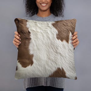 May include: A brown and white cowhide patterned throw pillow. The pillow is being held by a person.