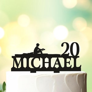 May include: Black cake topper with the name "MICHAEL" and the number "20". The topper features a silhouette of a person rowing a boat. The cake is white and decorated with greenery.