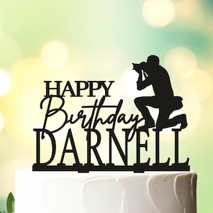 May include: Black acrylic cake topper with a silhouette of a photographer kneeling and taking a picture. The text "Happy Birthday Darnell" is written below the silhouette.