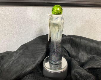 May include: A decorative bottle stopper with a green faceted glass orb on top. The ceramic stopper has a white, blue, and black design with glitter accents. It sits on a silver base, against a black textured background.