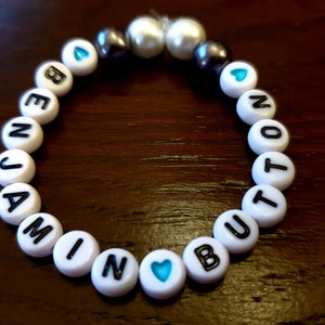 May include: A beaded bracelet with white letter beads spelling "BENJAMIN BUT NOT". The bracelet also features blue heart beads, pearl beads, and dark gray beads. The bracelet is on a dark brown wooden surface.