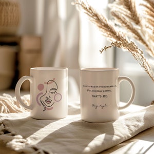 May include: Two white ceramic mugs with black text and designs. One mug features a line drawing of a woman's face with pink and purple circles. The other mug has the quote "I am a woman phenomenally. Phenomenal woman, that's me." by Maya Angelou.
