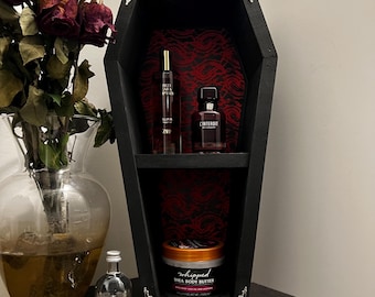 May include: A black coffin-shaped shelf with silver corner accents, holding perfume bottles and a jar of shea body butter. The shelf's interior features a red patterned fabric. A glass skull and a vase of dried roses are also present.
