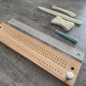 Puede incluir: Un conjunto de herramientas de artesanía, que incluye una tabla de clavijas de madera con pomos blancos, una regla transparente, un ganchillo con mango azul y plegaderas. Las herramientas están dispuestas sobre una superficie gris.