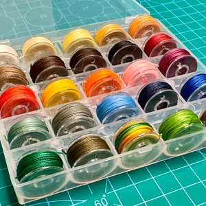 May include: A clear plastic storage box with 20 compartments filled with spools of colorful thread. The spools are arranged in rows and columns.