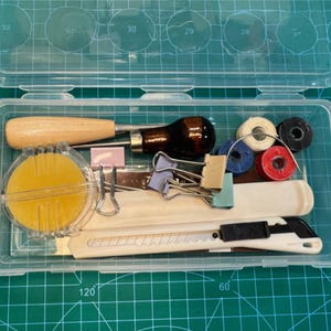 May include: A clear plastic craft kit box containing various tools. Includes a wooden handled awl, a dark brown handled tool, spools of thread, paper clips, a yellow sponge, a white utility knife, and other small items. The box is on a green cutting mat.
