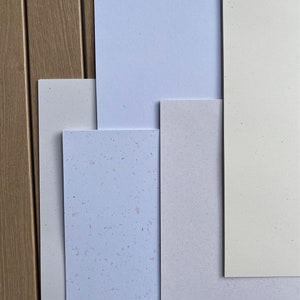 Op de afbeelding: Een verzameling van vijf verschillende papierstalen, waaronder een lichtblauwe, een witte, een lichtbeige, een beige met spikkels en een lichtbeige met spikkels. De stalen zijn in een verspringend patroon gerangschikt.