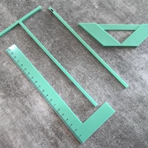 Puede incluir: Un conjunto de herramientas de dibujo de color verde menta, que incluye una regla con marcas de centímetros, una escuadra en T, un ángulo recto, un triángulo y una varilla larga y delgada. Las herramientas están dispuestas sobre una superficie gris texturizada.