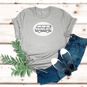 A light gray t-shirt lays flat. The shirt has a white circular embellishment featuring a graphic of a van that reads VAN LIFE