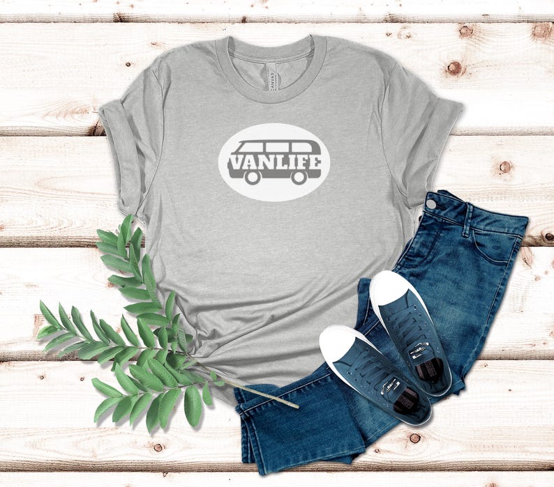 A light gray t-shirt lays flat. The shirt has a white circular embellishment featuring a graphic of a van that reads VAN LIFE