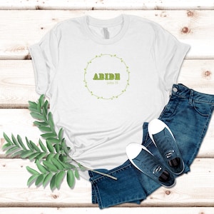 Puede incluir: Camiseta blanca con la palabra "ABIDE" en verde, rodeada por una corona de hojas verdes. El texto "John 15" está debajo. La camiseta se combina con vaqueros azules y zapatillas deportivas. Una ramita de hojas verdes está en primer plano.