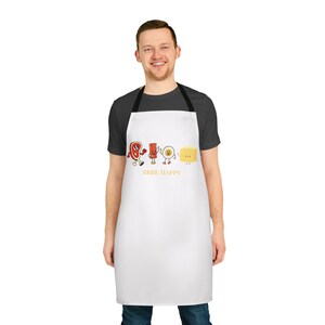 May include: White apron with a cartoon illustration of a steak, bacon, egg, and butter with the text "BRBE HAPPY". A person is wearing the apron.