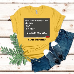 A yellow t-shirt that has a graphic of a blackboard on it. On the blackboard a lesson is written. It says, Believe in Yourselves, Dream, Try, Do Good. I Love You All. Class Dismissed.