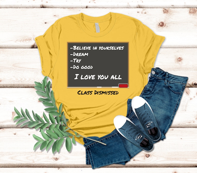 A yellow t-shirt that has a graphic of a blackboard on it. On the blackboard a lesson is written. It says, Believe in Yourselves, Dream, Try, Do Good. I Love You All. Class Dismissed.