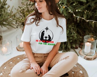 May include: White t-shirt featuring a black silhouette of the Nativity scene inside a circle, with the text "HE IS THE REASON FOR THE SEASON" in red and green. The shirt is worn by a person.