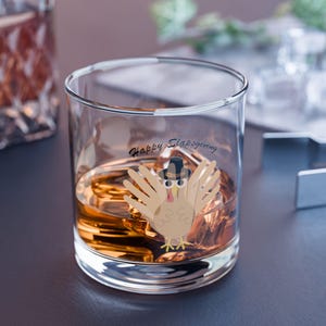 May include: A clear glass tumbler filled with amber liquid and ice. The glass has a cartoon turkey wearing a hat, with the text "Happy Slapsgiving". The glass is on a dark surface.