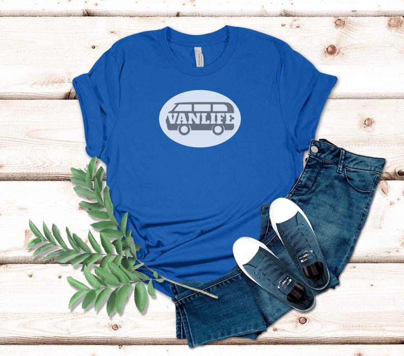 A blue t-shirt lays flat. The shirt has a white circular embellishment featuring a graphic of a van that reads VAN LIFE