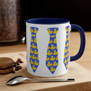 May include: White ceramic mug with a blue rim and handle. The mug features a design of three blue ties made of yellow rubber duckies.