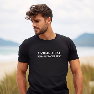 May include: Black crew neck t-shirt with the text "A STEAK A DAY KEEPS THE DOCTOR AWAY" in white block letters. The shirt is made of a soft material and is suitable for casual wear.
