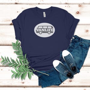A navy blue t-shirt lays flat. The shirt has a white circular embellishment featuring a graphic of a van that reads VAN LIFE
