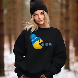 May include: Black sweatshirt with a yellow Pac-Man character wearing a blue and white winter hat and eating snowflakes.