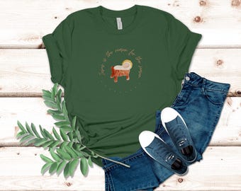 May include: A dark green t-shirt with the text "Jesus is the reason for the season" encircling a watercolor illustration of a manger. The shirt is paired with blue jeans and dark blue sneakers.