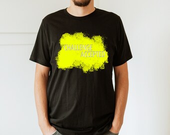 Puede incluir: Camiseta negra con el texto "CHALLENGE ACCEPTED" en letras blancas sobre una forma amarilla brillante texturizada. La camiseta es de estilo cuello redondo.
