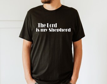 Może przedstawiać: Czarny t-shirt z białym napisem "The Lord is my Shepherd". Koszulka ma okrągły dekolt i wydaje się być wykonana z miękkiego materiału. Osoba ma na sobie szare dżinsy.
