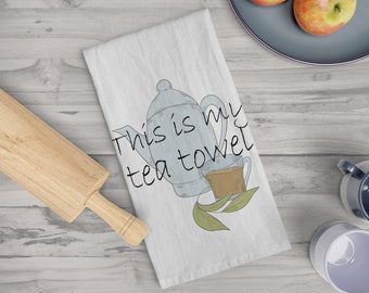 May include: White tea towel with a blue teapot and teacup illustration. The text "This is my tea towel" is written on the towel. A wooden rolling pin and two apples on a plate are also visible.